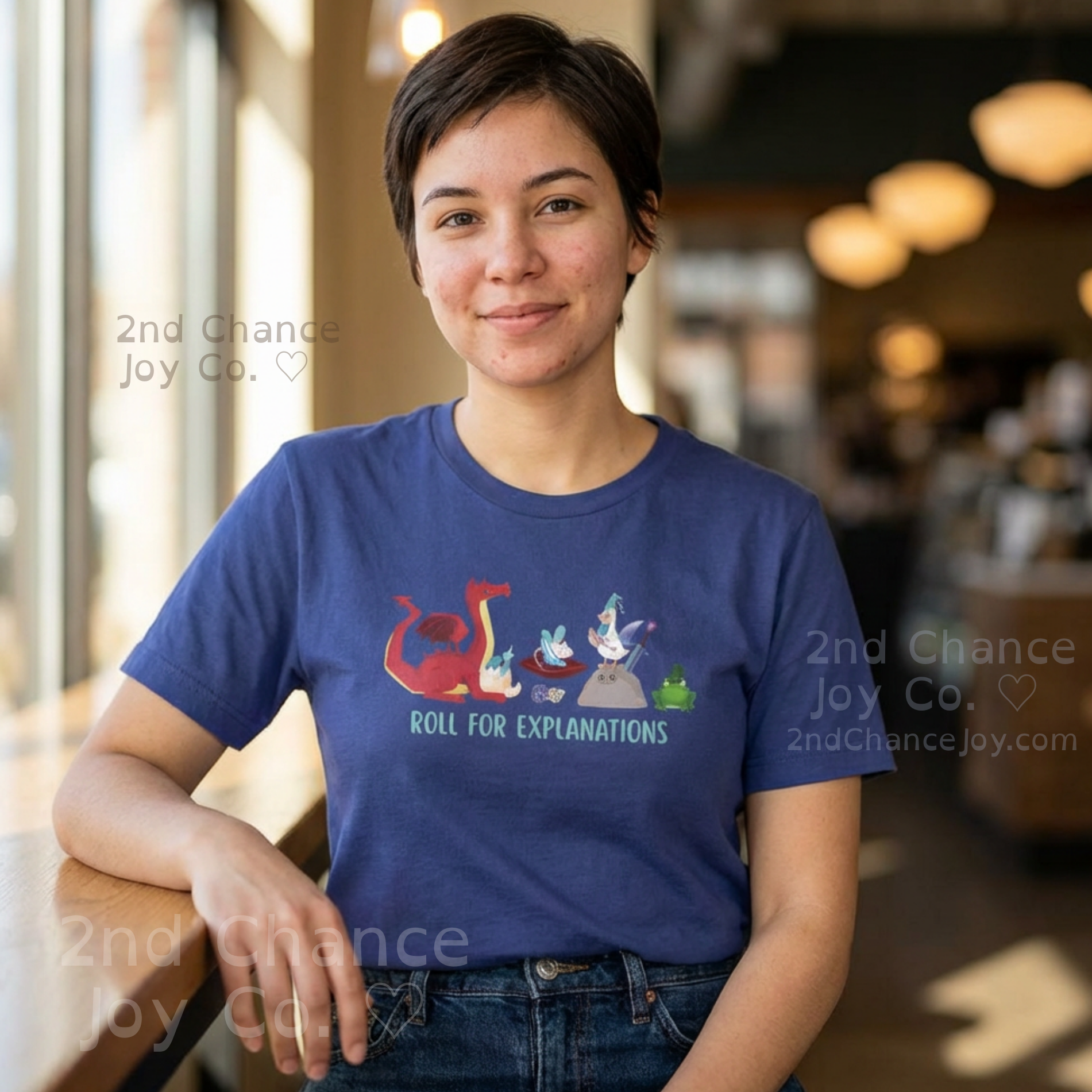 Woman with brown pixie-cut hair wearing  a blue t-shirt with fantasy roleplaying game inspired characters and the text Roll for explanations". She sits in a casual setting